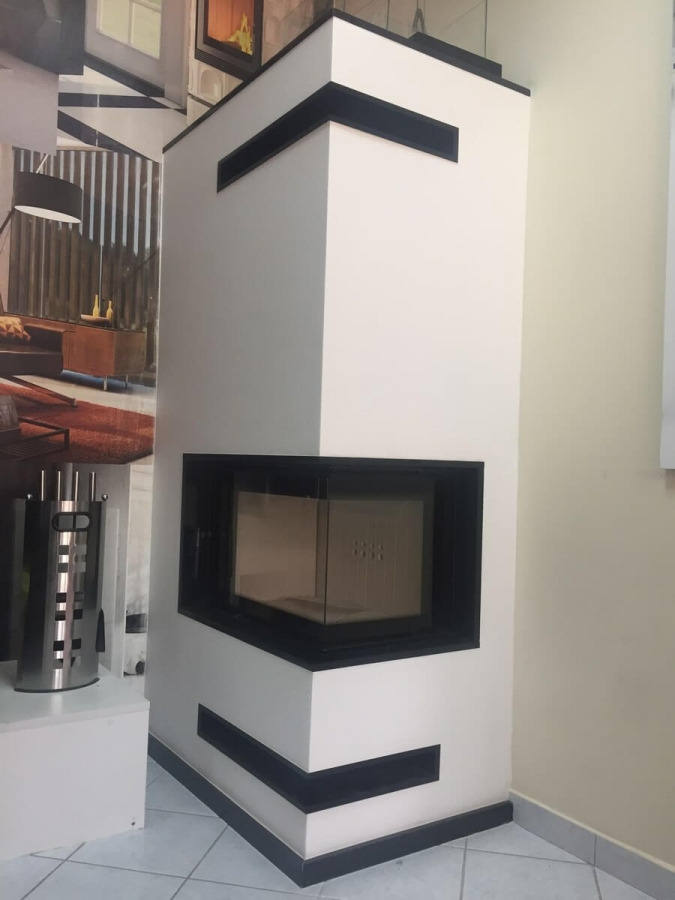 Biały, narożny wkład kominkowy z czarnymi akcentami, widoczne akcesoria kominkowe i aranżacja wnętrza w tle.