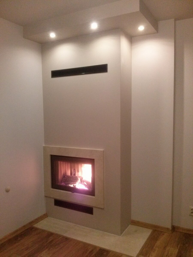 Nowoczesny, wnękowy palenisko z płomieniem, obudowane beżowym kamieniem, wbudowane w ścianę z minimalistycznym, czarnym grillem wentylacyjnym u góry.