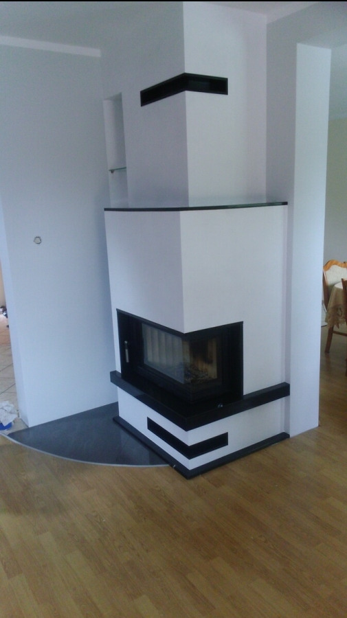 Nowoczesny, narożny wkład kominkowy z czarnym obramowaniem i białym wykończeniem, umieszczony na czarnej, półokrągłej płycie, z widocznym paleniskiem i drewnianą podłogą w tle.