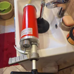 Czerwony pneumatyczny przepychacz do rur z manometrem i wężem, ustawiony w białej umywalce obok chromowanej baterii i gumowego przepychacza. Widoczne mydło i kosmetyki. Niska perspektywa...