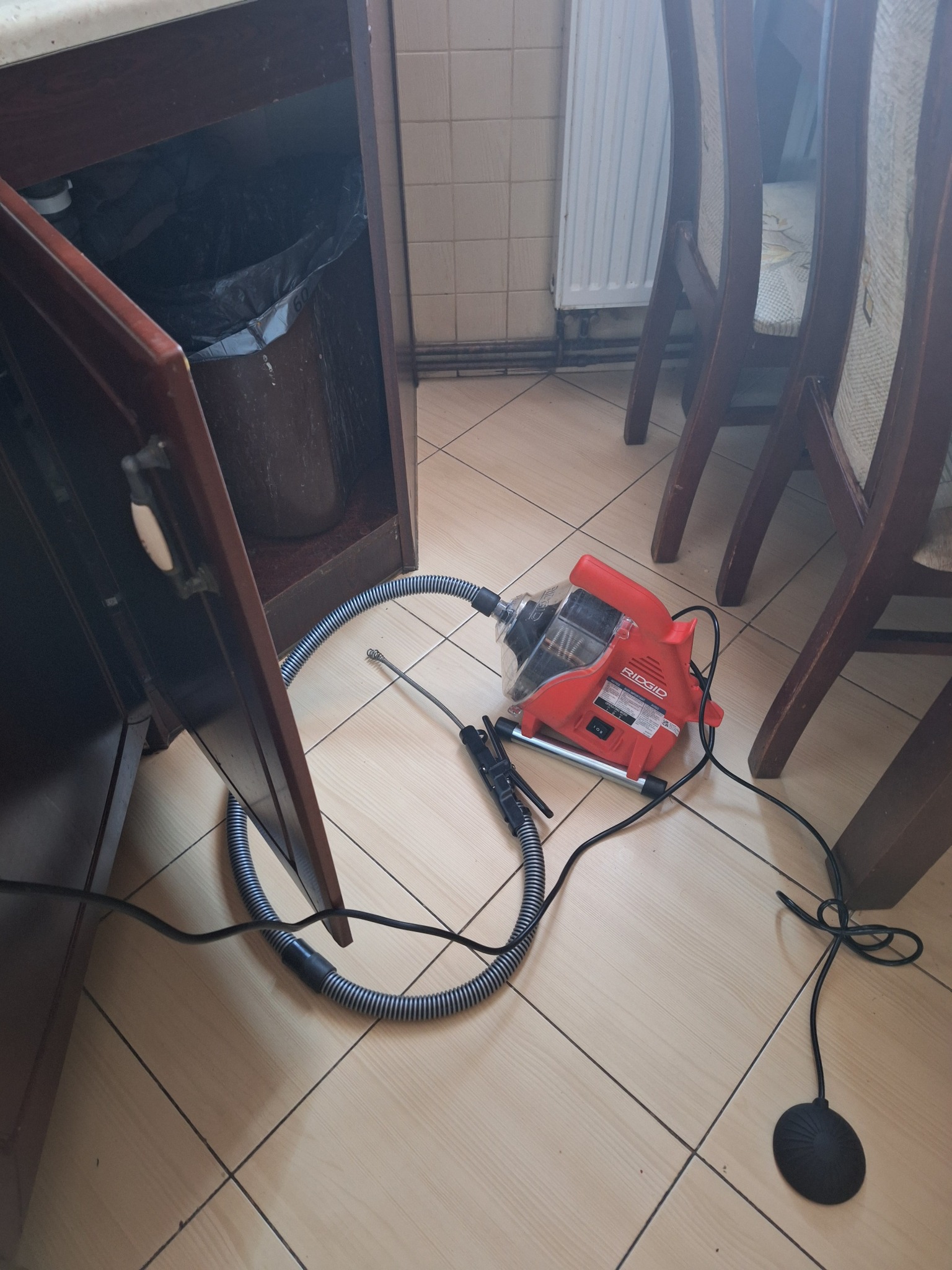 Czerwona elektryczna przepychaczka Ridgid leży na podłodze z płytek obok otwartej szafki zlewozmywakowej z widocznym koszem na śmieci w środku, z podłączonym przewodem wchodzącym do odpływu.