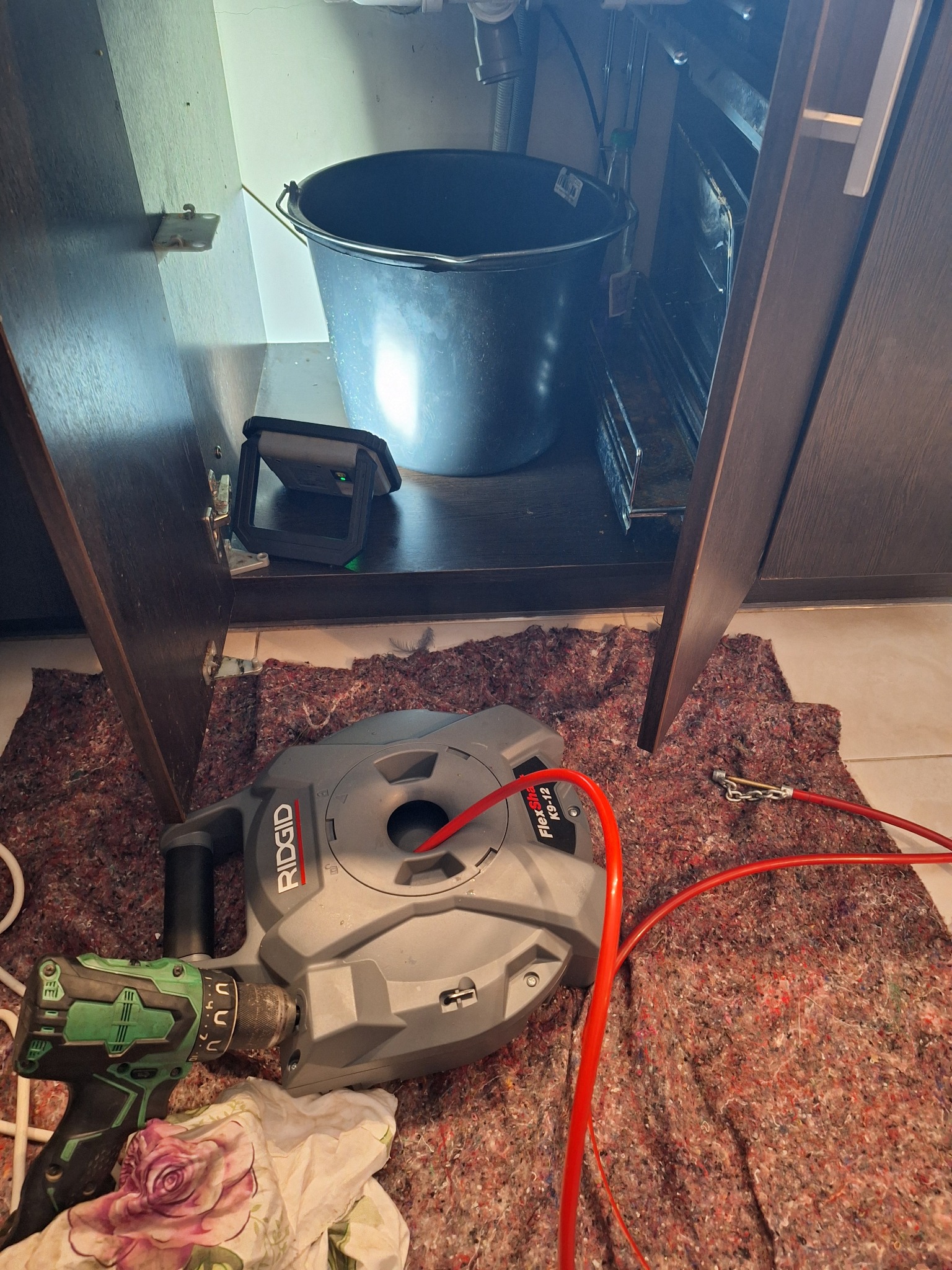 Otwarta szafka pod zlewem, widoczne wiadro, lampa warsztatowa i maszyna do czyszczenia rur Ridgid FlexShaft K9-12 z podłączoną wiertarką akumulatorową na brudnej szmacie.