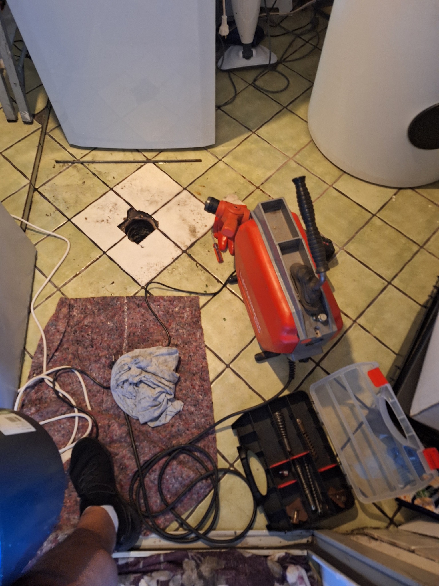 Otwór w podłodze z widoczną kanalizacją, obok czerwona maszyna do przepychania rur, skrzynka z końcówkami, szmata i noga w czarnym bucie.