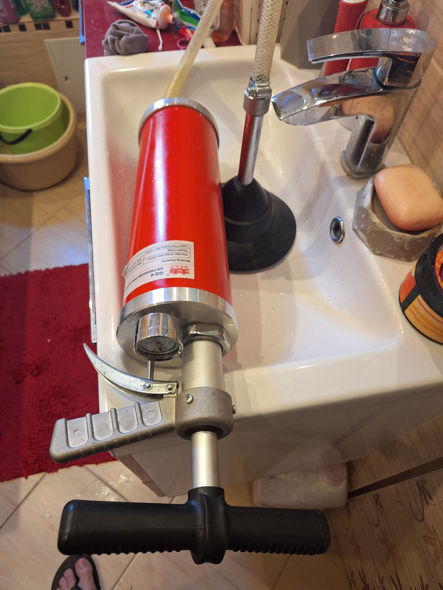 Czerwony pneumatyczny przepychacz do rur z manometrem i wężem, ustawiony w białej umywalce obok chromowanej baterii i gumowego przepychacza. Widoczne mydło i kosmetyki. Niska perspektywa...