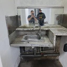 Kapitoshka - Zabrudzona metalowa umywalka w pomieszczeniu po remoncie, widoczne rury, ściany z resztkami farby, odbicie osoby robiącej zdjęcie w lustrze.