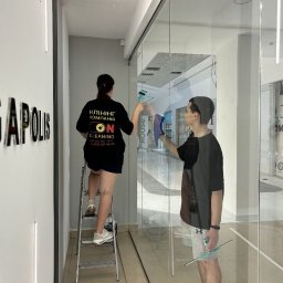 Kapitoshka - Dwie osoby czyszczą szklaną witrynę sklepową w galerii handlowej. Kobieta stoi na drabinie, mężczyzna używa ściągaczki do szyb.