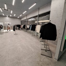 Kapitoshka - Wnętrze sklepu odzieżowego z szarymi ścianami, marmurową podłogą i rzędami ubrań na wieszakach, oświetlone podłużnymi lampami sufitowymi.