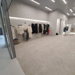 Kapitoshka - Wnętrze sklepu odzieżowego z jasnymi ścianami o fakturze betonu, metalową kratownicą sufitową i rzędami ubrań na wieszakach w odcieniach bieli i czerni, widok na marmurową podłogę.