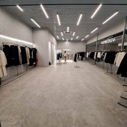 Kapitoshka - Wnętrze sklepu odzieżowego z marmurową posadzką, minimalistycznym wystrojem, oświetleniem LED i ubraniami na wieszakach po obu stronach.