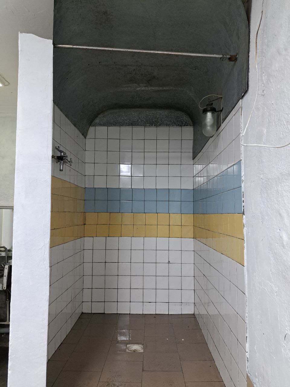 Wnętrze starej, wykafelkowanej kabiny prysznicowej z żółto-niebieskim pasem, widoczny odpływ w podłodze i lampa na ścianie.