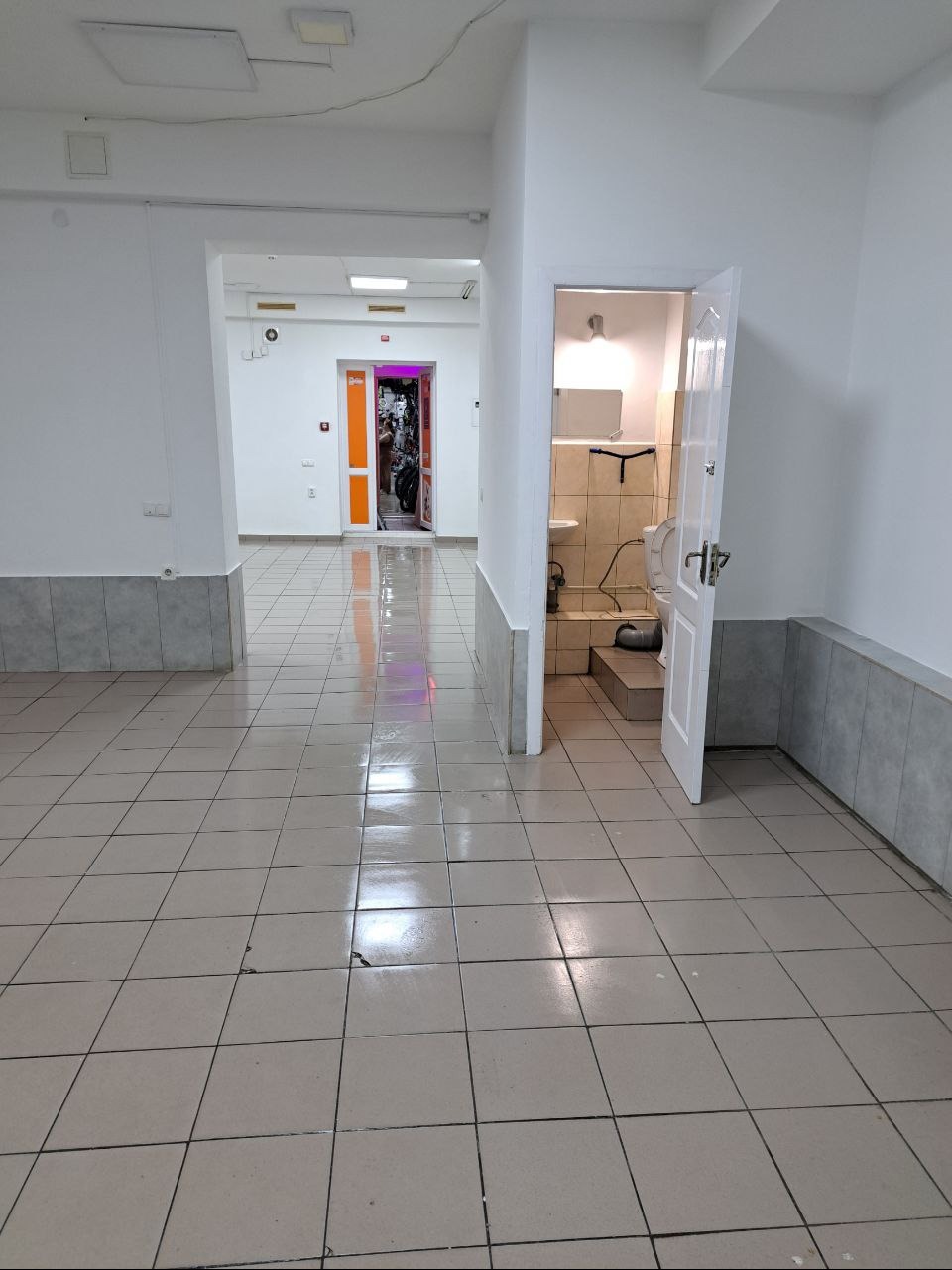 Pusta przestrzeń biurowa z widoczną toaletą, szara płytka podłogowa, białe ściany z szarym cokołem, otwarte drzwi do pomieszczenia z rowerami w tle.