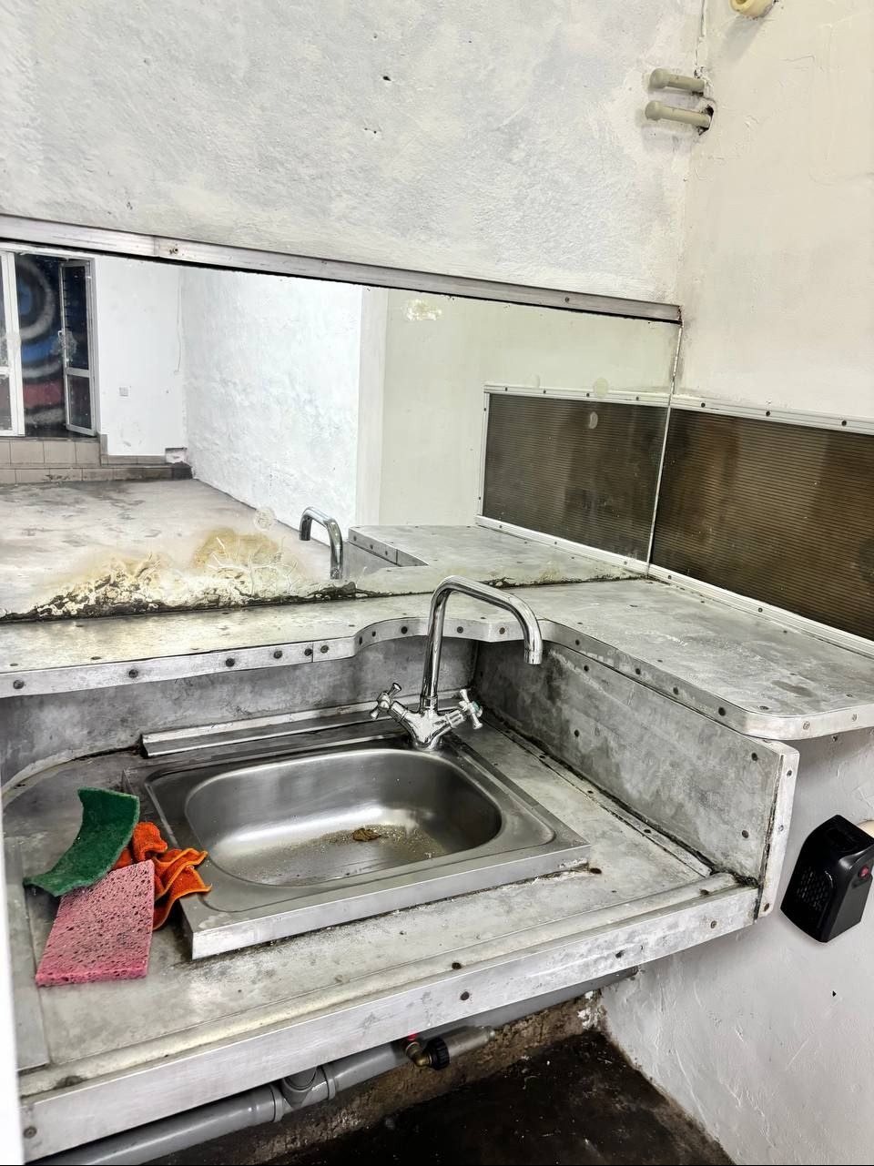 Zaniedbane pomieszczenie z metalową umywalką, widoczne gąbki do czyszczenia, brudne blaty i podłoga, odbicie w lustrze ukazuje dalszą część zniszczonego wnętrza.