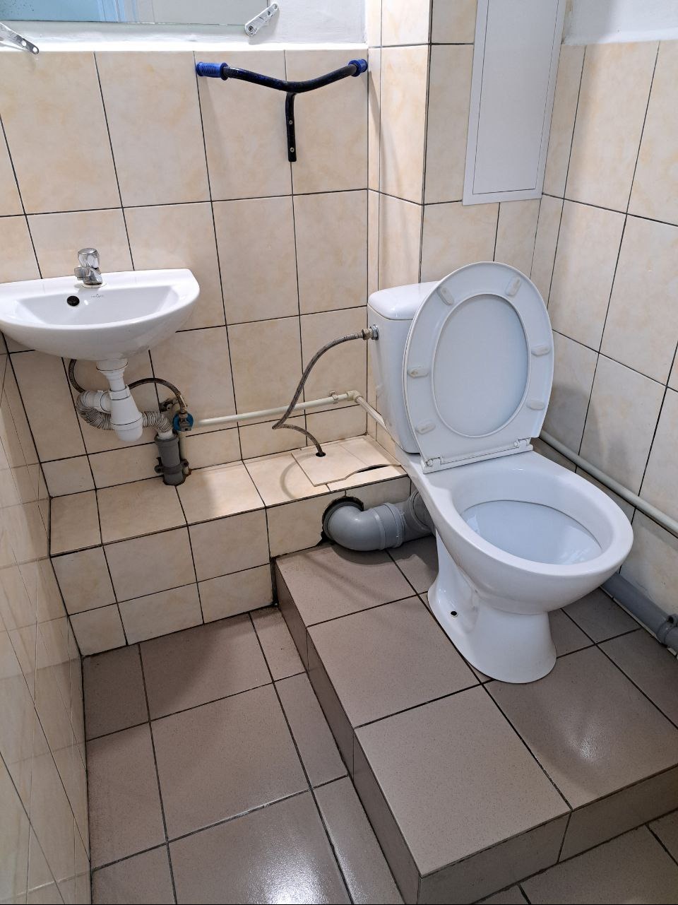 Standardowa toaleta z umywalką i muszlą klozetową w małej łazience wyłożonej beżowymi płytkami ceramicznymi, widoczne rury i uchwyt dla niepełnosprawnych.