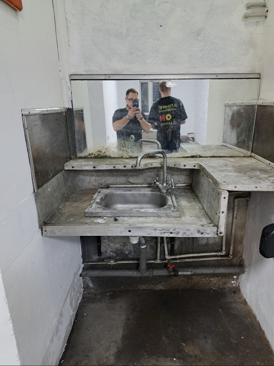 Zabrudzona metalowa umywalka w pomieszczeniu po remoncie, widoczne rury, ściany z resztkami farby, odbicie osoby robiącej zdjęcie w lustrze.