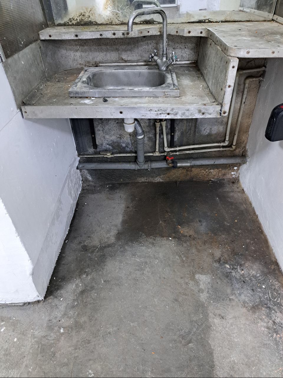 Zaniedbana, metalowa umywalka z widocznymi rurami i zabrudzonym otoczeniem, w tym mokra i brudna podłoga betonowa.