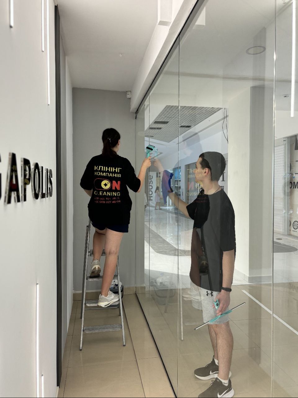 Dwie osoby czyszczą szklaną witrynę sklepową w galerii handlowej. Kobieta stoi na drabinie, mężczyzna używa ściągaczki do szyb.