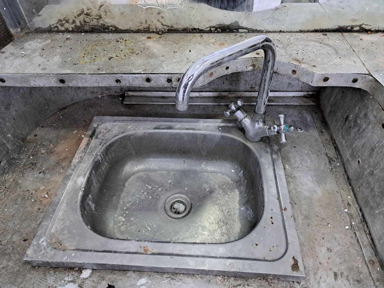 Zabrudzony, metalowy zlew z chromowaną baterią i śladami rdzy, otoczony resztkami gruzu i zanieczyszczeń, sugerujący potrzebę intensywnego czyszczenia.