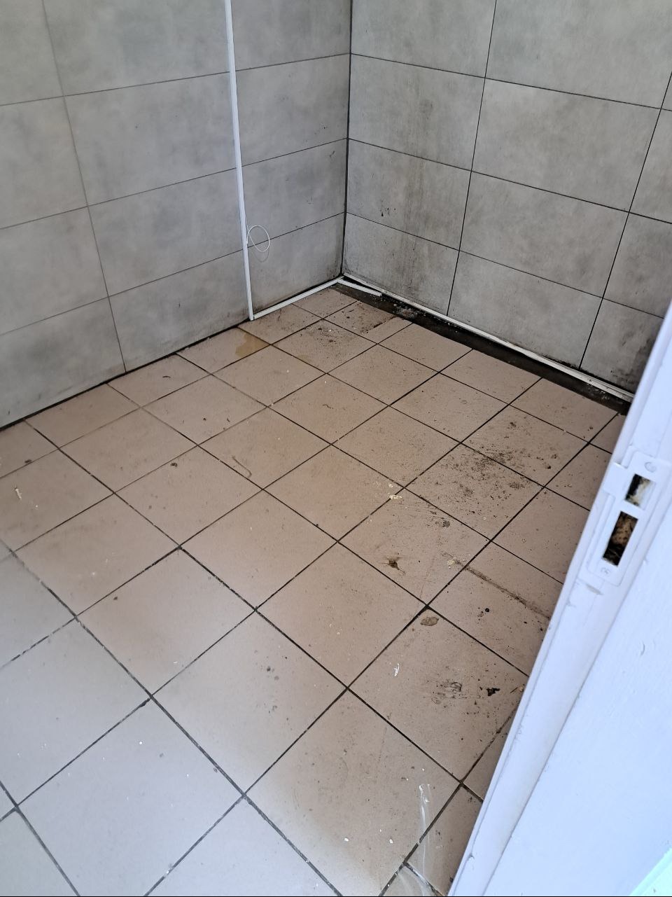 Brudna, kafelkowana podłoga i ściany w rogu pomieszczenia, widoczne plamy i zabrudzenia przed sprzątaniem.