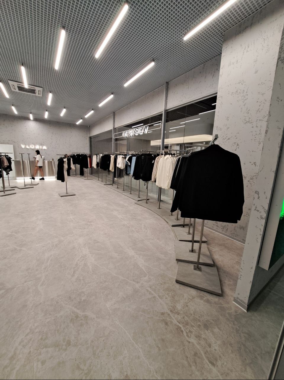 Wnętrze sklepu odzieżowego z szarymi ścianami, marmurową podłogą i rzędami ubrań na wieszakach, oświetlone podłużnymi lampami sufitowymi.