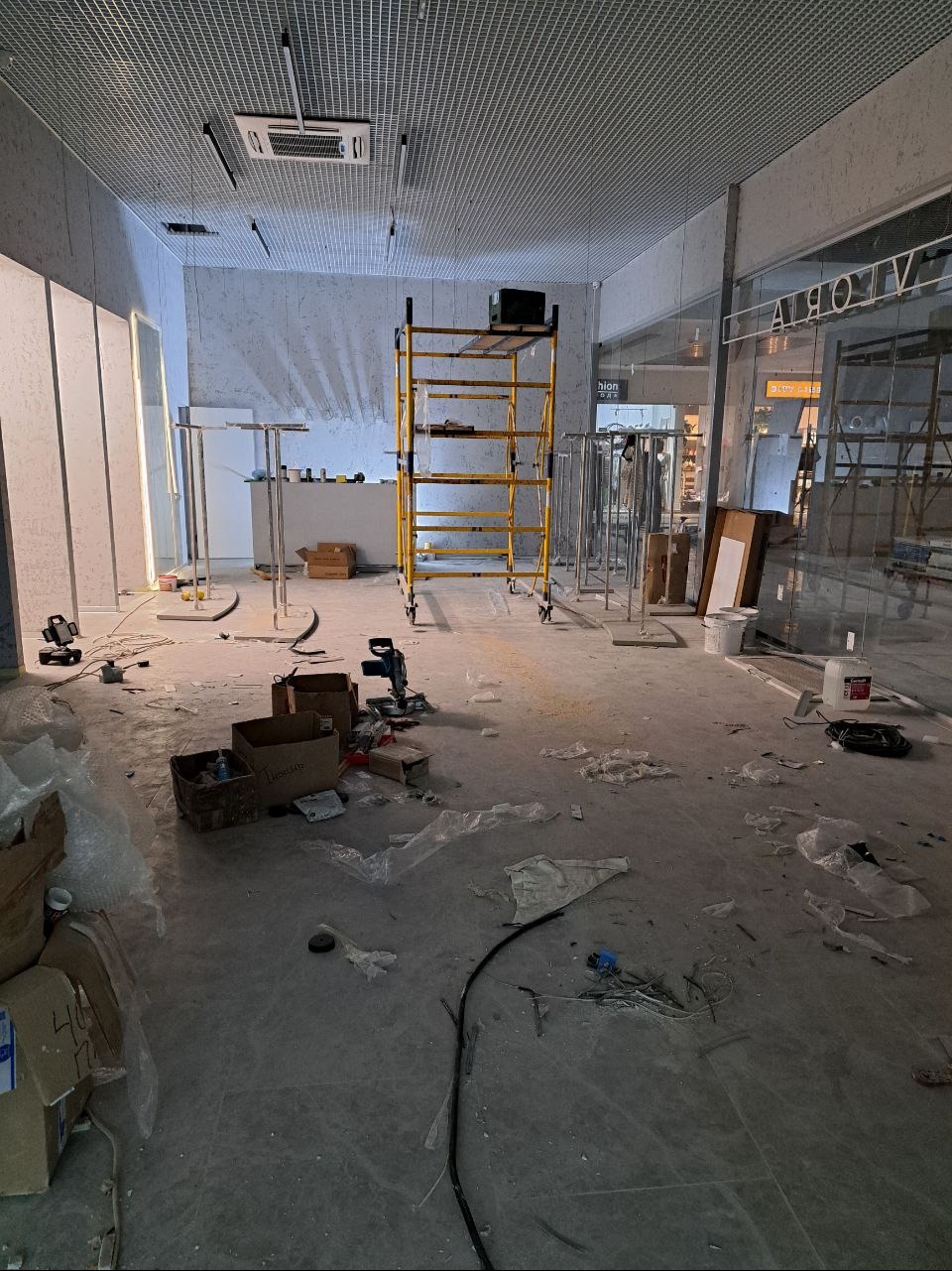 Wnętrze sklepu w trakcie prac wykończeniowych: żółte rusztowanie, rozłożone narzędzia, kartony i folie na podłodze.