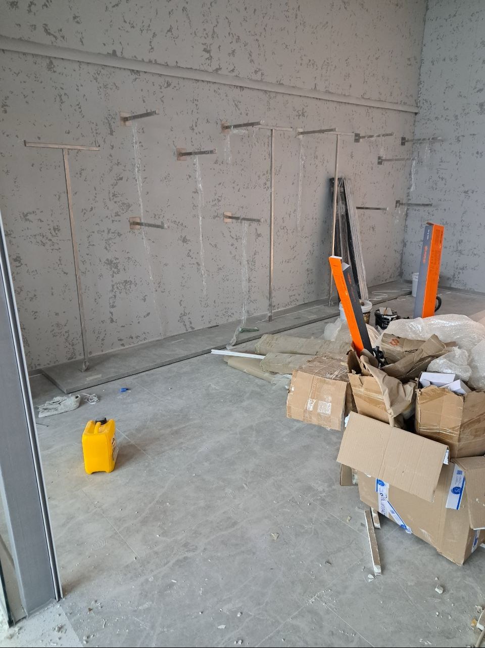 Wnętrze lokalu w trakcie prac wykończeniowych; widoczne metalowe uchwyty na ścianie, kartony, wiaderko z żółtą farbą i resztki materiałów budowlanych na podłodze.