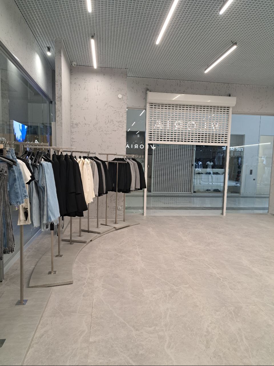 Jasne, minimalistyczne wnętrze sklepu odzieżowego z ubraniami na wieszakach i zamkniętą roletą antywłamaniową.