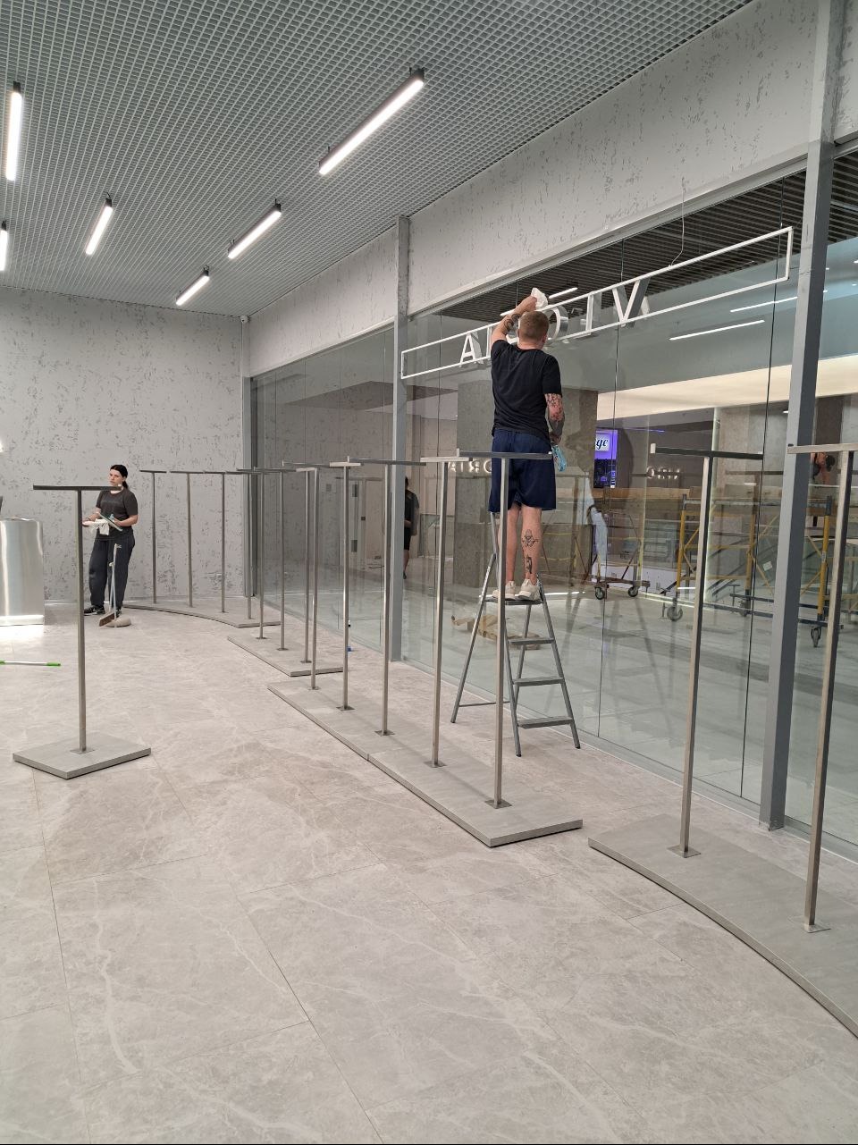 Pracownik na drabinie czyści szklaną witrynę sklepową z napisem, drugi pracownik w tle, widoczne metalowe stojaki na ubrania.