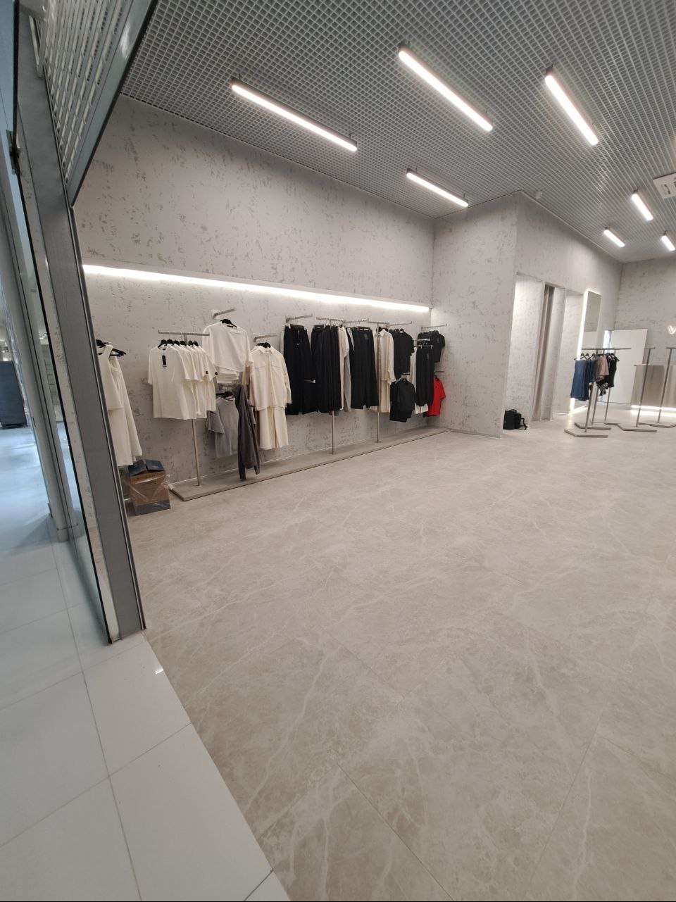 Wnętrze sklepu odzieżowego z jasnymi ścianami o fakturze betonu, metalową kratownicą sufitową i rzędami ubrań na wieszakach w odcieniach bieli i czerni, widok na marmurową podłogę.