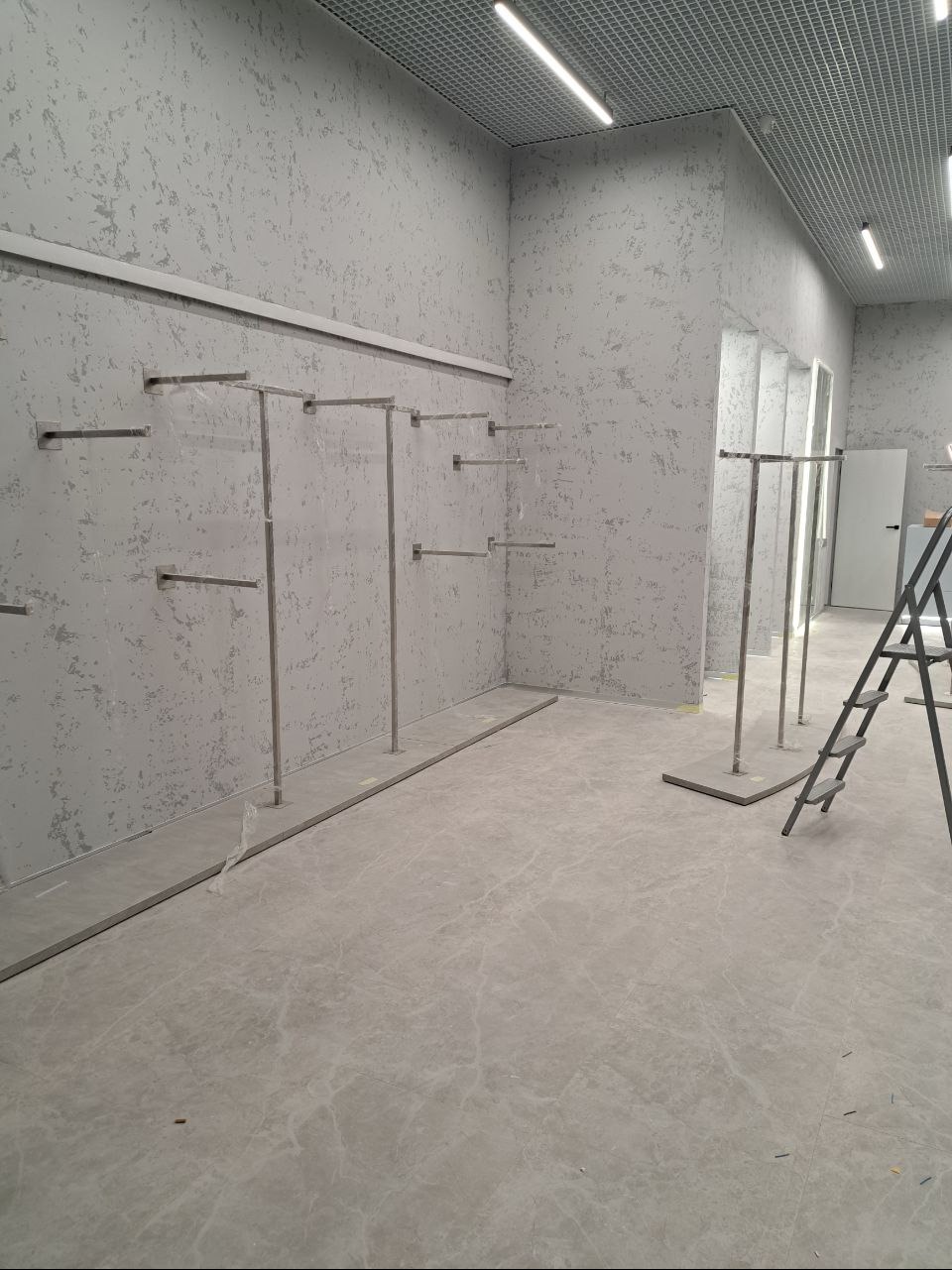 Pusty lokal handlowy po remoncie: szare ściany z fakturą, metalowe wieszaki na ubrania zamontowane na ścianach, drabina oparta o ścianę, szara marmurowa podłoga, sufit rastrowy z oświetleniem.