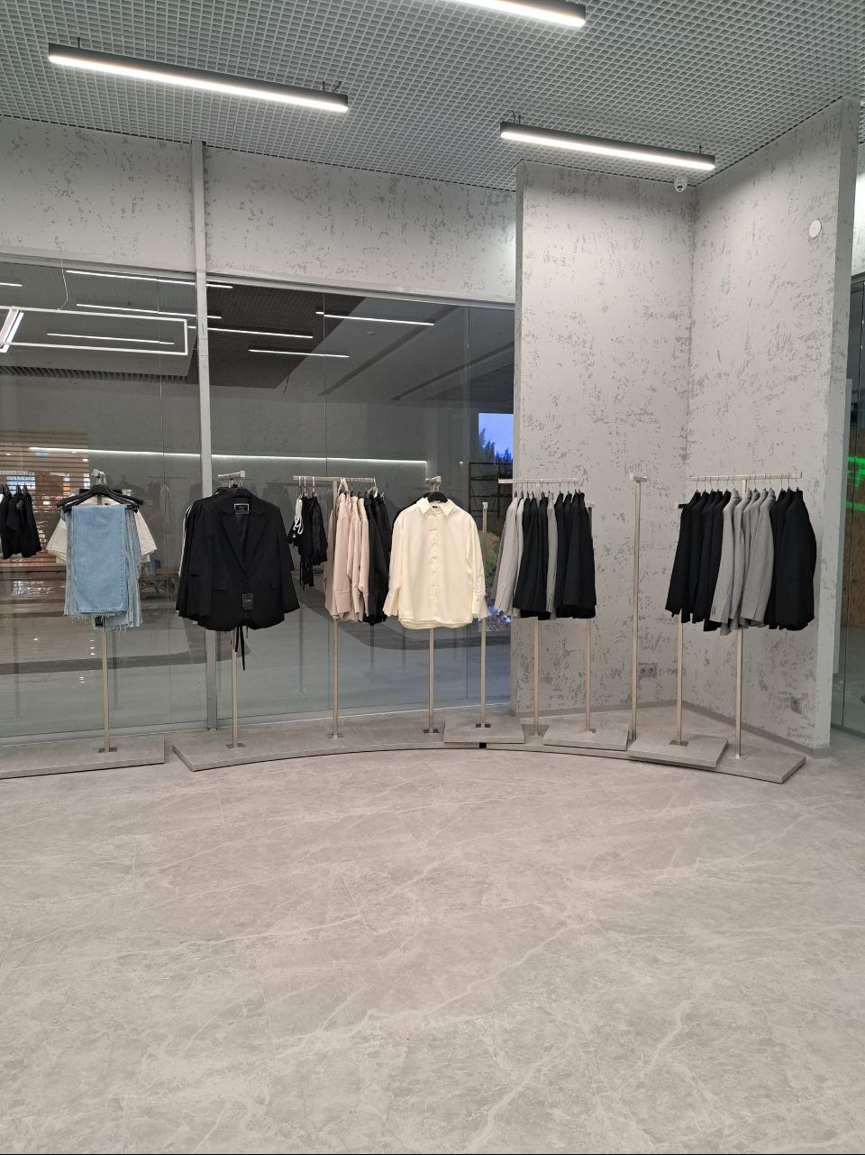 Minimalistyczny wystrój sklepu odzieżowego z ubraniami zawieszonymi na metalowych wieszakach na tle szarej marmurowej podłogi i teksturowanej ściany.