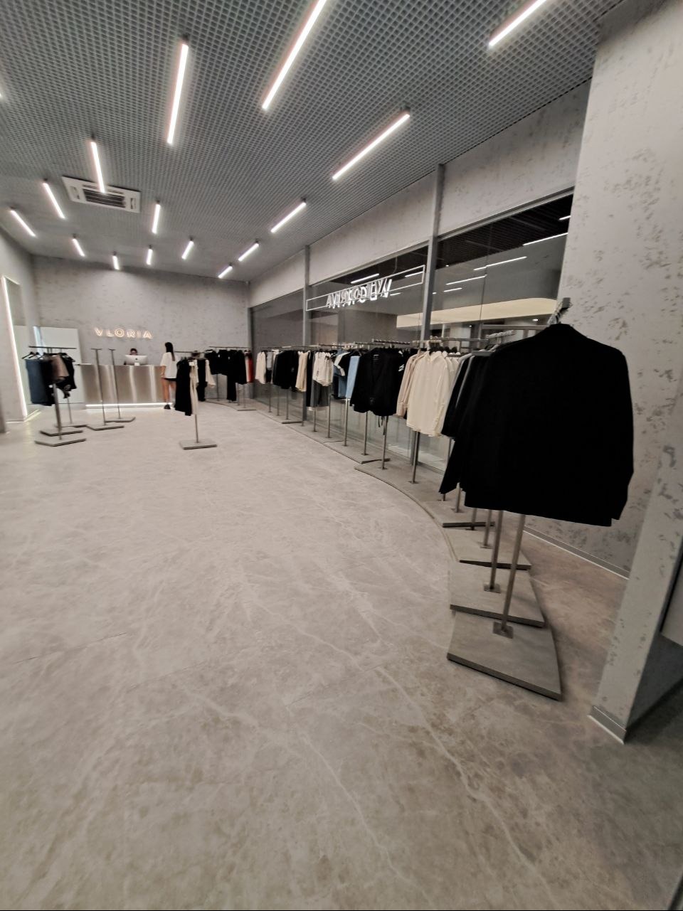 Przestronne wnętrze butiku odzieżowego z marmurową podłogą, oświetleniem ledowym i minimalistycznym wystrojem, prezentujące kolekcję ubrań na wieszakach.