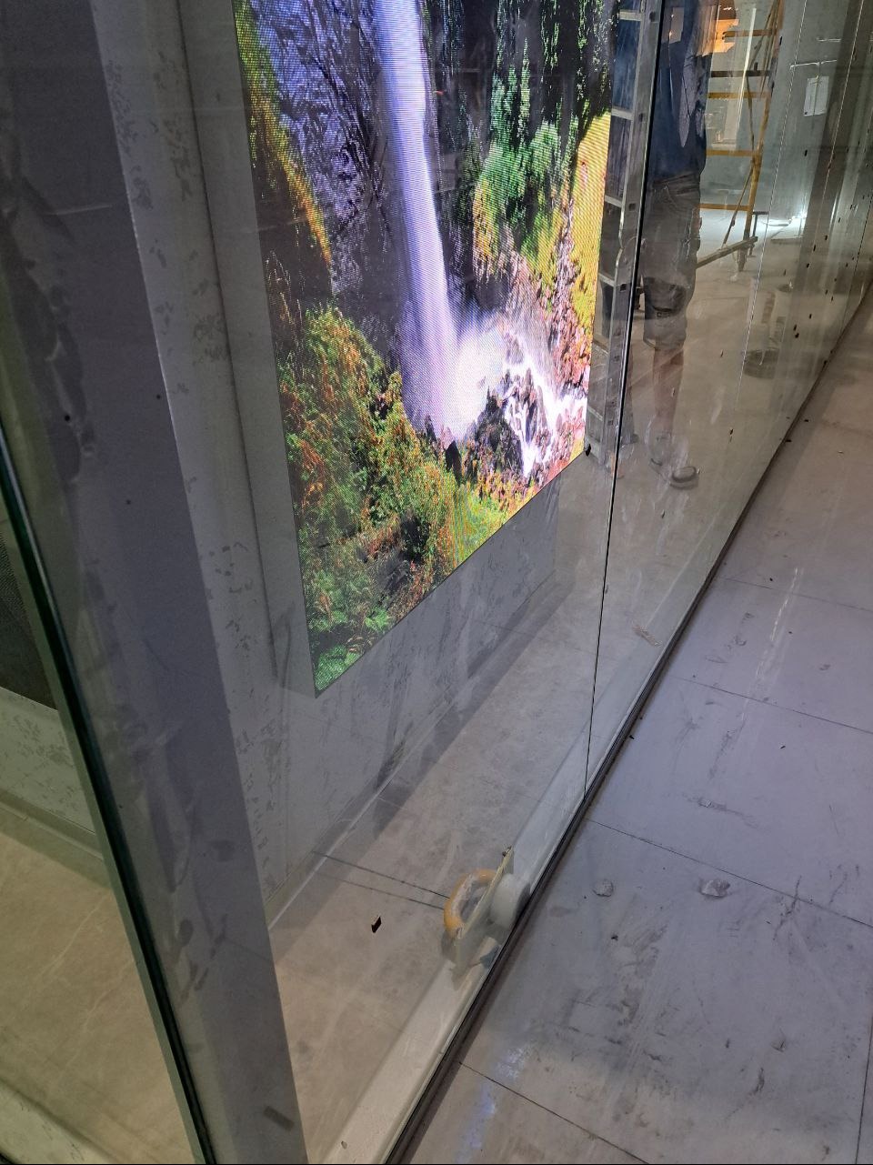 Montaż szklanej ściany z wbudowanym ekranem LED prezentującym wodospad; widoczne ślady po budowie na marmurowej posadzce i osoba na drabinie w tle.