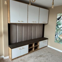 Claro Adrian Popiołek - Zabudowa wnęki z szafkami górnymi, tapicerowanym panelem ściennym i siedziskiem z szafkami dolnymi w stylu safari, oświetlona żarówkami dekoracyjnymi na linach jutowych.