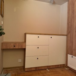 Claro Adrian Popiołek - Zabudowa wnęki z szafkami i blatem z szufladą, fronty w kolorze złamanej bieli, korpus i blat w odcieniu drewna, widoczne gniazdko elektryczne i fragment parkietu.