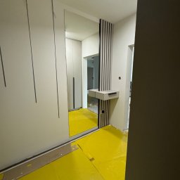 Claro Adrian Popiołek - Wnętrze w trakcie wykańczania: widoczna szafa z lustrem i pionowymi wgłębieniami na frontach, obok konsola z pionowymi listwami dekoracyjnymi, podłoga zabezpieczona folią ochronną.