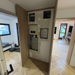 Claro Adrian Popiołek - Narożna szafka z drewnianymi drzwiczkami maskująca instalację elektryczną i skrzynkę rozdzielczą w trakcie montażu, widoczne niedoskonałości wykończenia ściany.