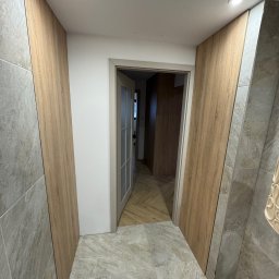 Claro Adrian Popiołek - Korytarz z zabudową ścienną z paneli drewnopodobnych, szara podłoga imitująca kamień, otwarte drzwi do kolejnego pomieszczenia z podłogą w jodełkę.