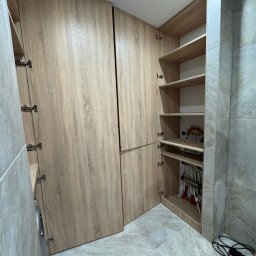Claro Adrian Popiołek - Wysoka zabudowa z szaf z frontami z jasnego drewna, z otwartymi drzwiczkami ukazującymi półki i instalację hydrauliczną wewnątrz, na tle ściany wyłożonej płytkami imitującymi kamień.