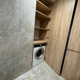 Claro Adrian Popiołek - Pralka w zabudowie z drewnianymi frontami, otwarte półki nad pralką, szare płytki na ścianie i podłodze.