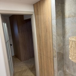 Claro Adrian Popiołek - Wnętrze łazienki z szafką z drewnopodobną okleiną, widok na szafę wnękową w korytarzu, szara płytka imitująca kamień na ścianie, lampa z ażurowym abażurem.