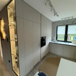 Nowoczesna kuchnia z zabudową w odcieniach szarości, podświetlana witryna z kieliszkami, minimalistyczne oświetlenie sufitowe i marmurowe wykończenie ściany przy oknie.