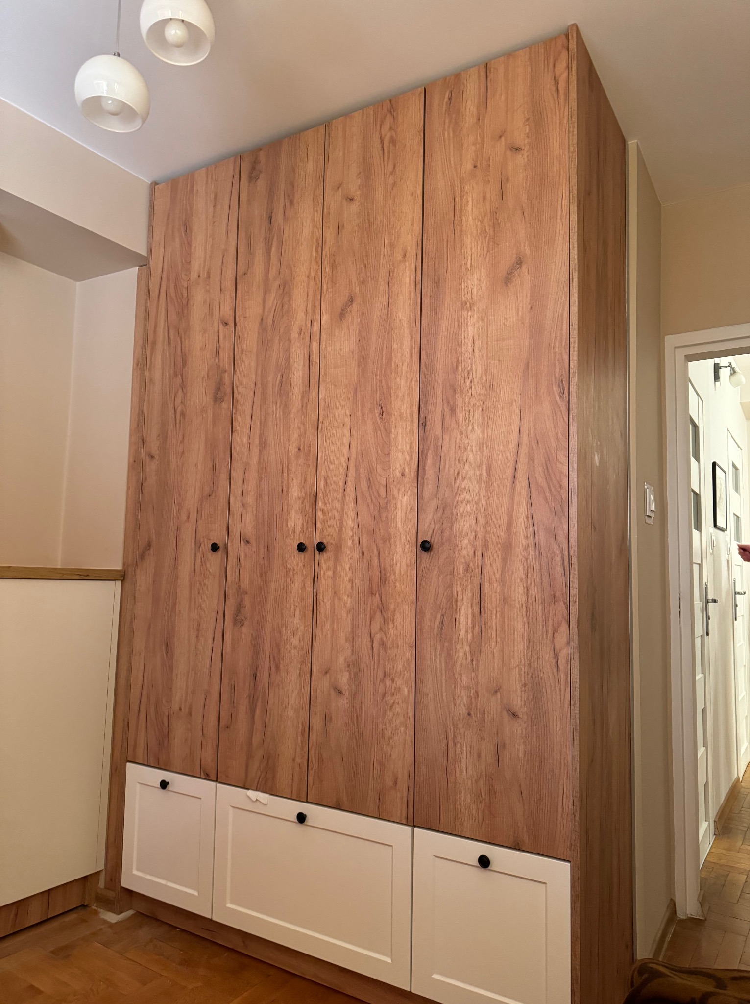 Wysoka, drewnopodobna szafa z białymi dolnymi szafkami i czarnymi gałkami, oświetlona trzema kulistymi lampami sufitowymi, widziana z perspektywy podłogi w mieszkaniu.