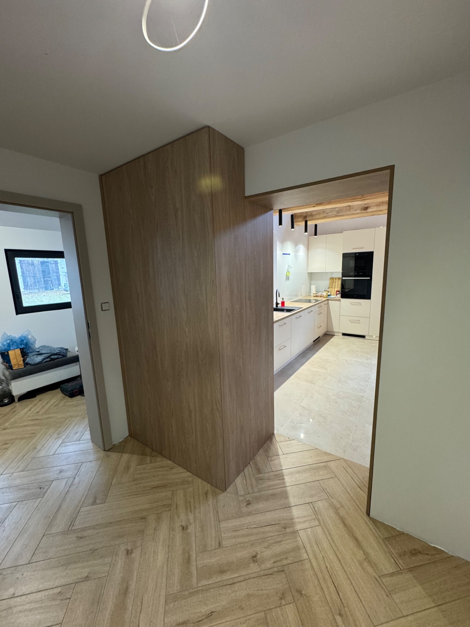 Nowoczesna kuchnia z zabudową meblową w odcieniach bieli, widziana z korytarza z szafą wnękową o drewnianym wykończeniu i podłogą w jodełkę.