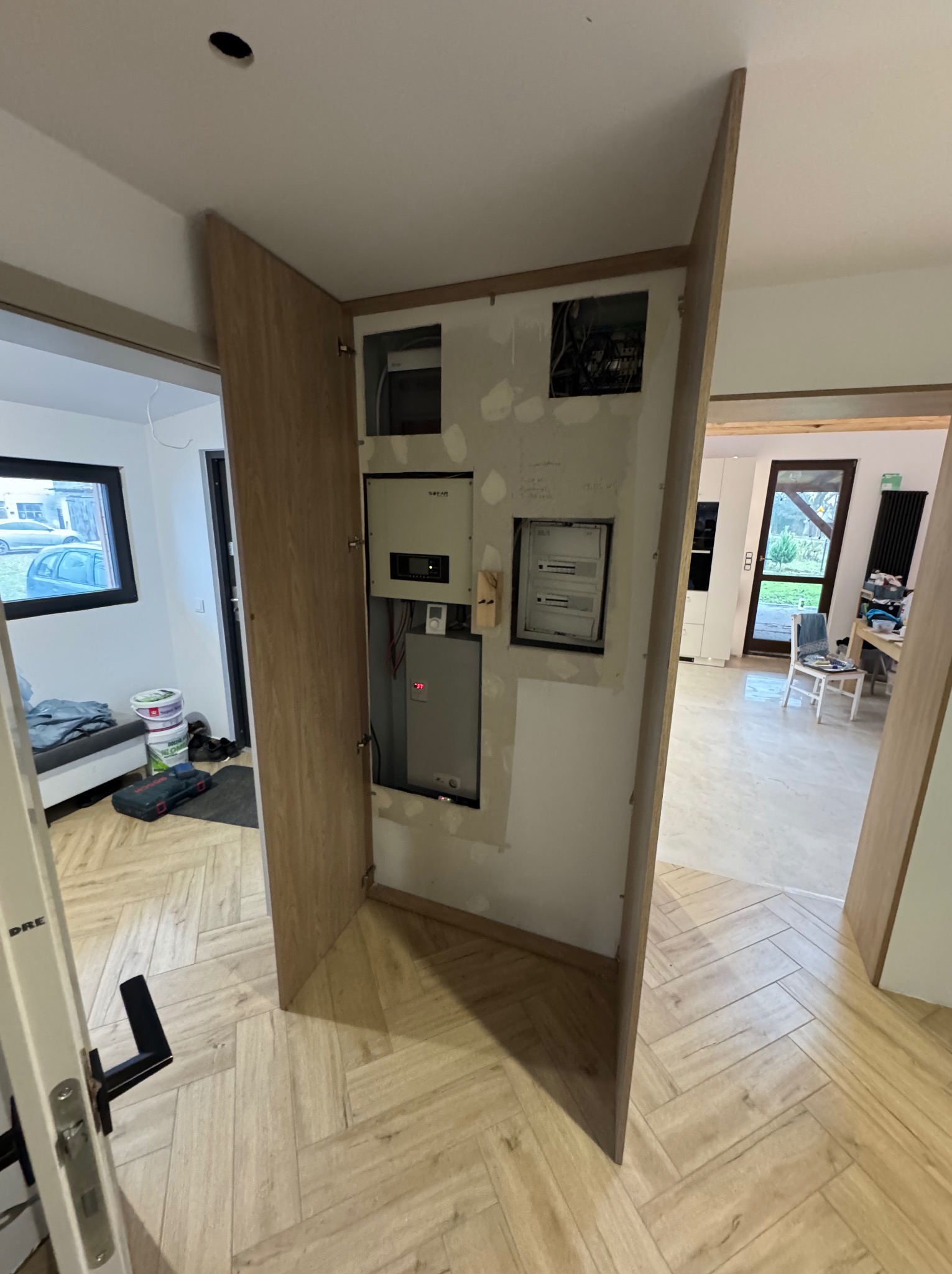 Narożna szafka z drewnianymi drzwiczkami maskująca instalację elektryczną i skrzynkę rozdzielczą w trakcie montażu, widoczne niedoskonałości wykończenia ściany.