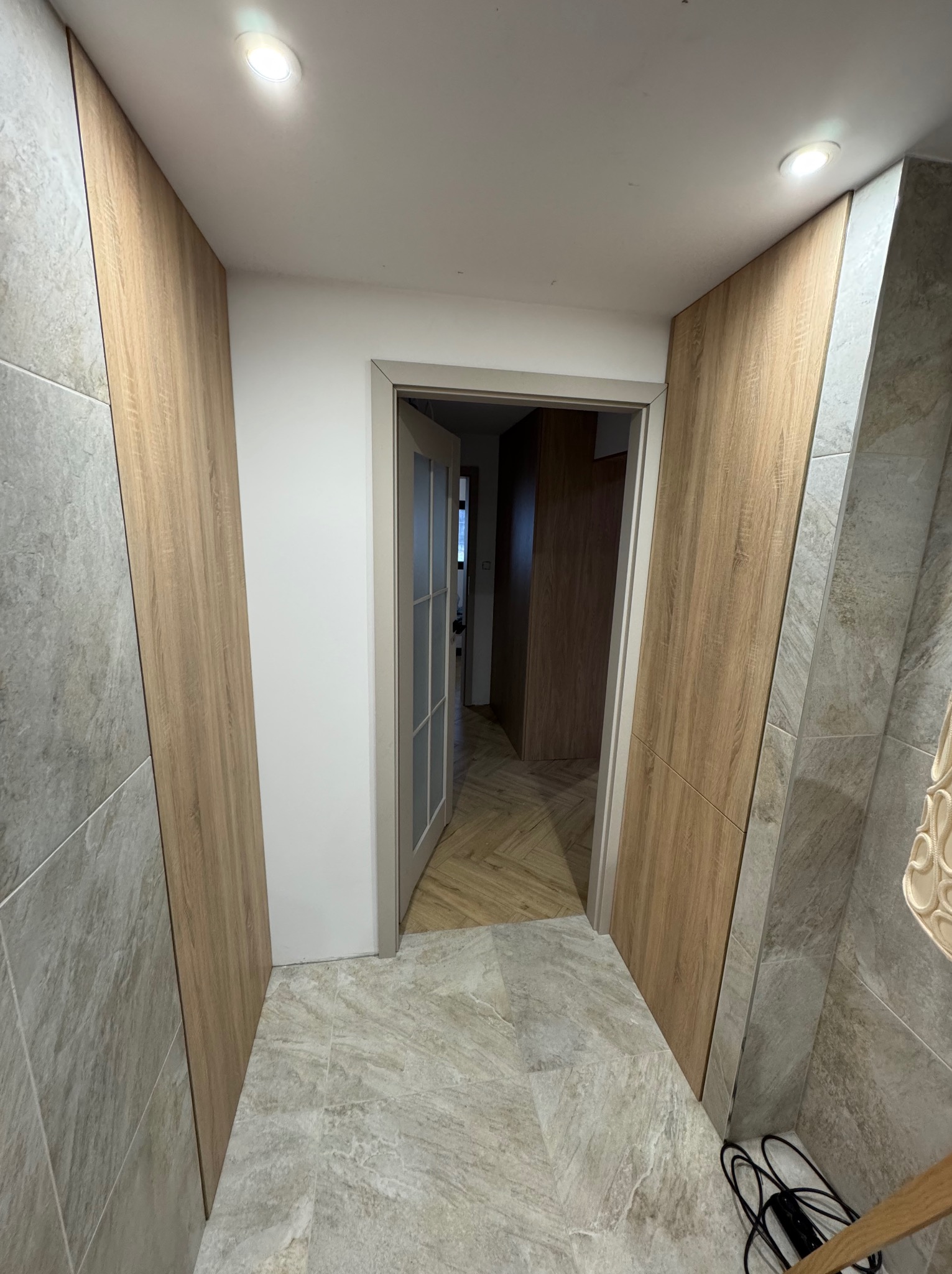 Korytarz z zabudową ścienną z paneli drewnopodobnych, szara podłoga imitująca kamień, otwarte drzwi do kolejnego pomieszczenia z podłogą w jodełkę.