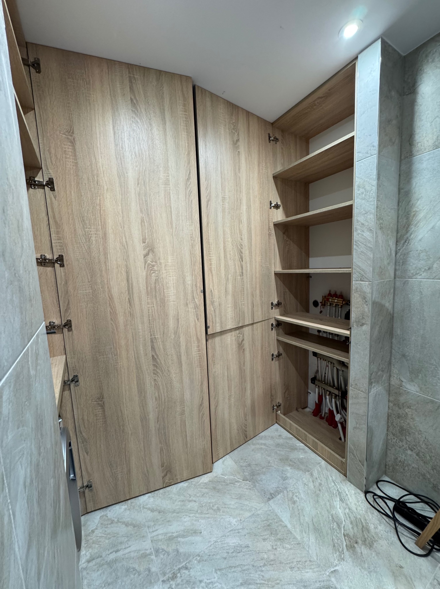 Wysoka zabudowa z szaf z frontami z jasnego drewna, z otwartymi drzwiczkami ukazującymi półki i instalację hydrauliczną wewnątrz, na tle ściany wyłożonej płytkami imitującymi kamień.