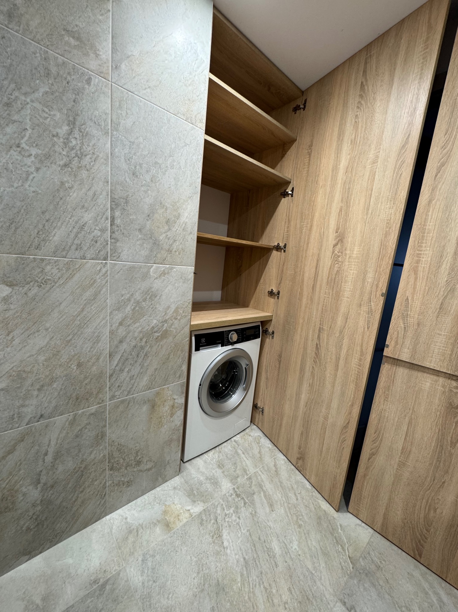 Pralka w zabudowie z drewnianymi frontami, otwarte półki nad pralką, szare płytki na ścianie i podłodze.