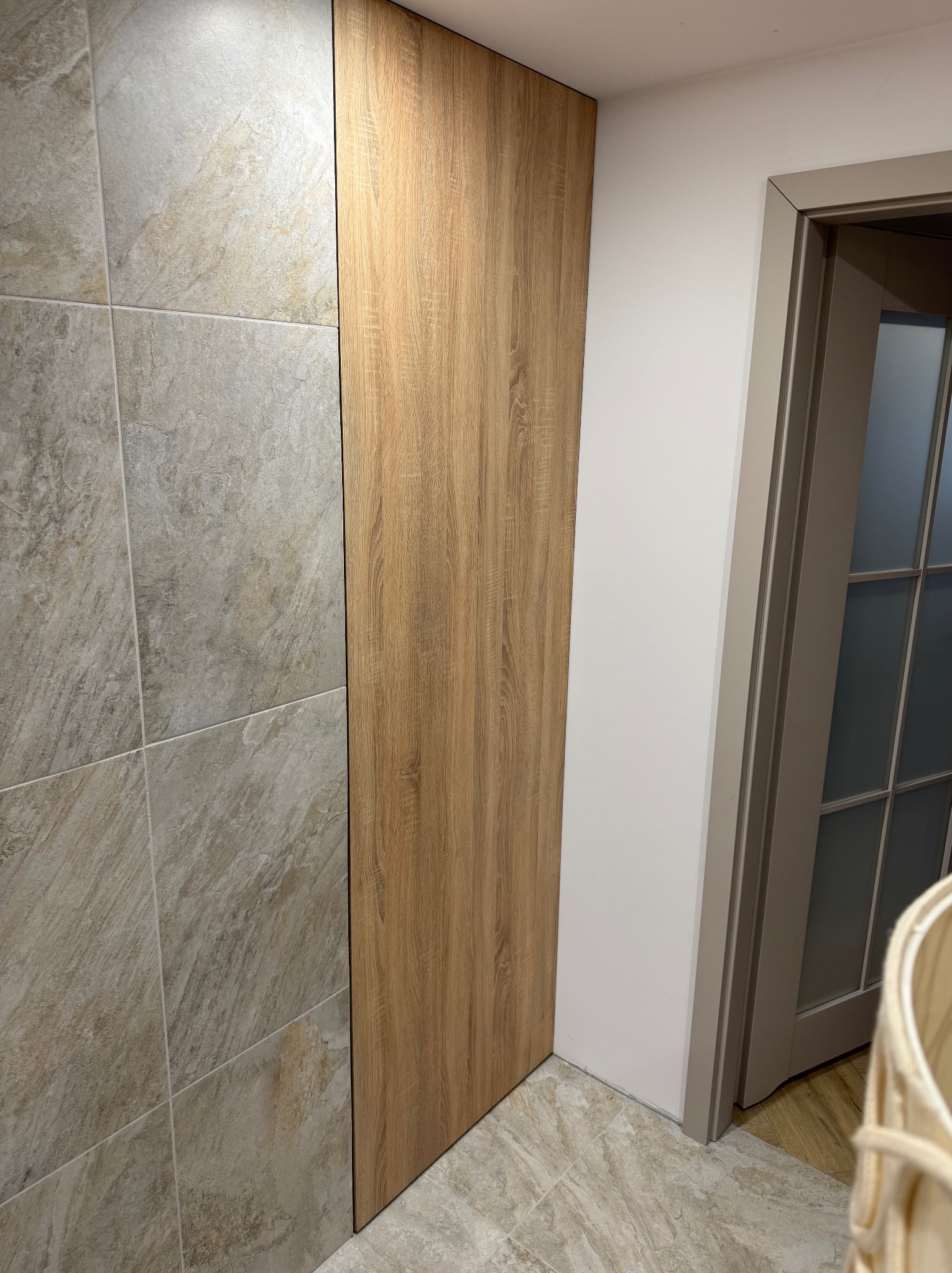 Pionowa zabudowa z drewnopodobnej płyty w przedpokoju, sąsiadująca z płytkami imitującymi kamień, obok ościeżnica drzwiowa w odcieniach szarości.