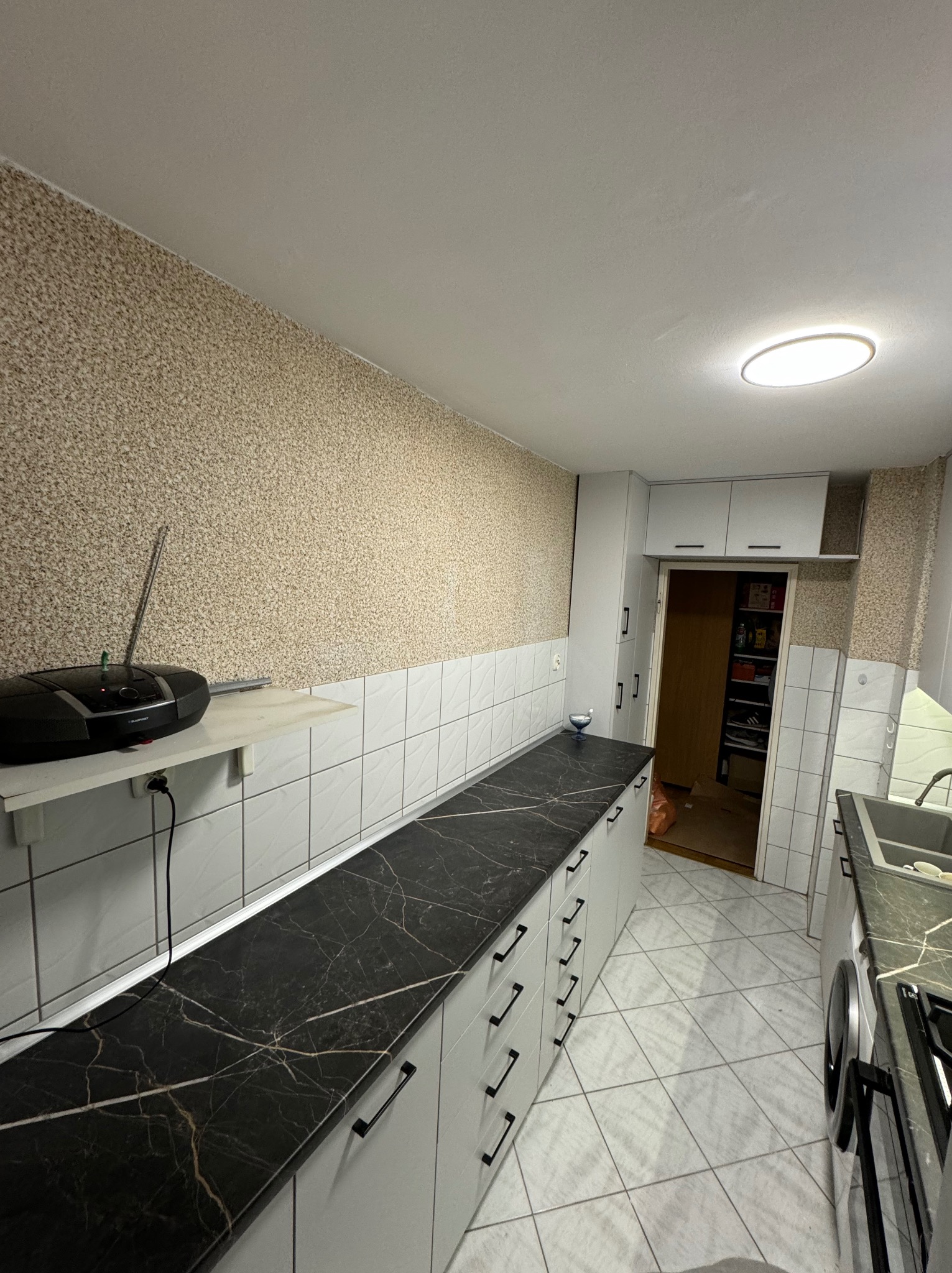 Długa, wąska kuchnia z białymi szafkami i czarnym blatem imitującym marmur, widoczny odtwarzacz CD na półce i wejście do spiżarni.