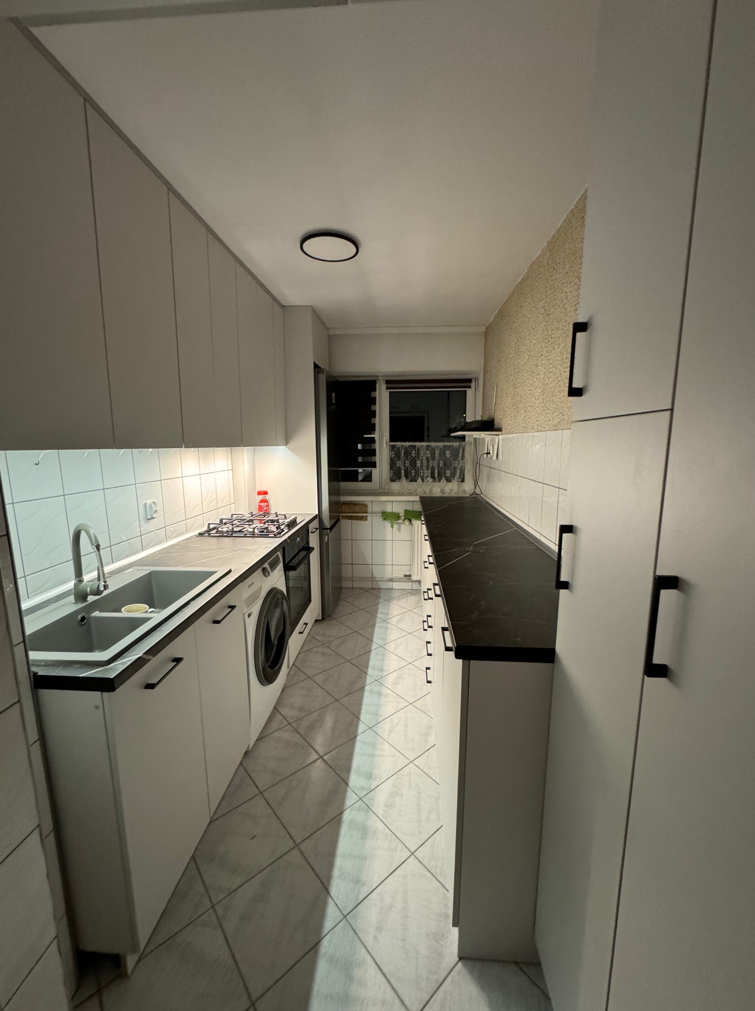 Wąska kuchnia z białymi szafkami, czarnym blatem i szarą podłogą w romby; pralka w zabudowie obok zlewu i płyty gazowej.